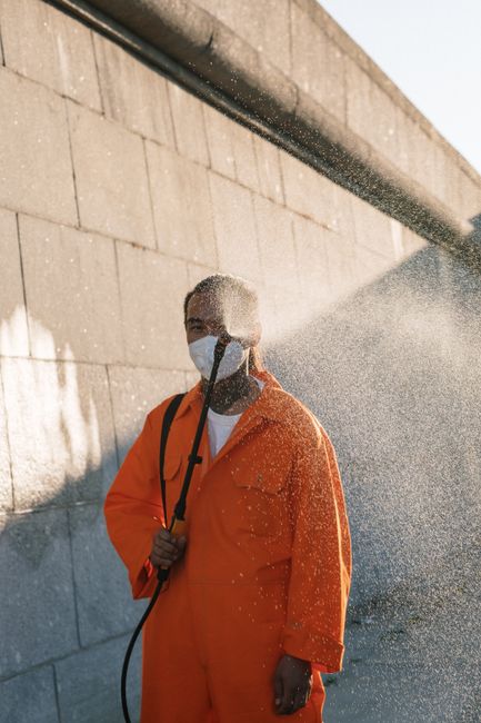 A person wearing an orange waterproof protective suit and a face mask is using a pressure washer to clean the exterior concrete wall of a building. The high-pressure water spray creates a mist and vis