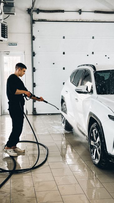 A person wearing an orange waterproof protective suit and a face mask is using a pressure washer to clean the exterior concrete wall of a building. The high-pressure water spray creates a mist and vis
