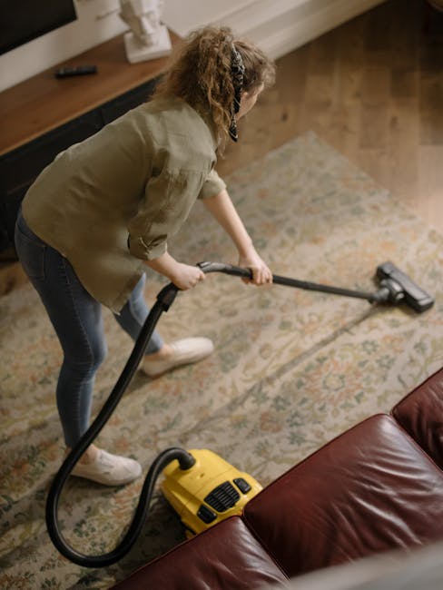A person kneeling on a patterned rug in a residential room, using a yellow portable vacuum cleaner with a black hose to deep clean the carpet. The individual is wearing casual clothing, with a beige j
